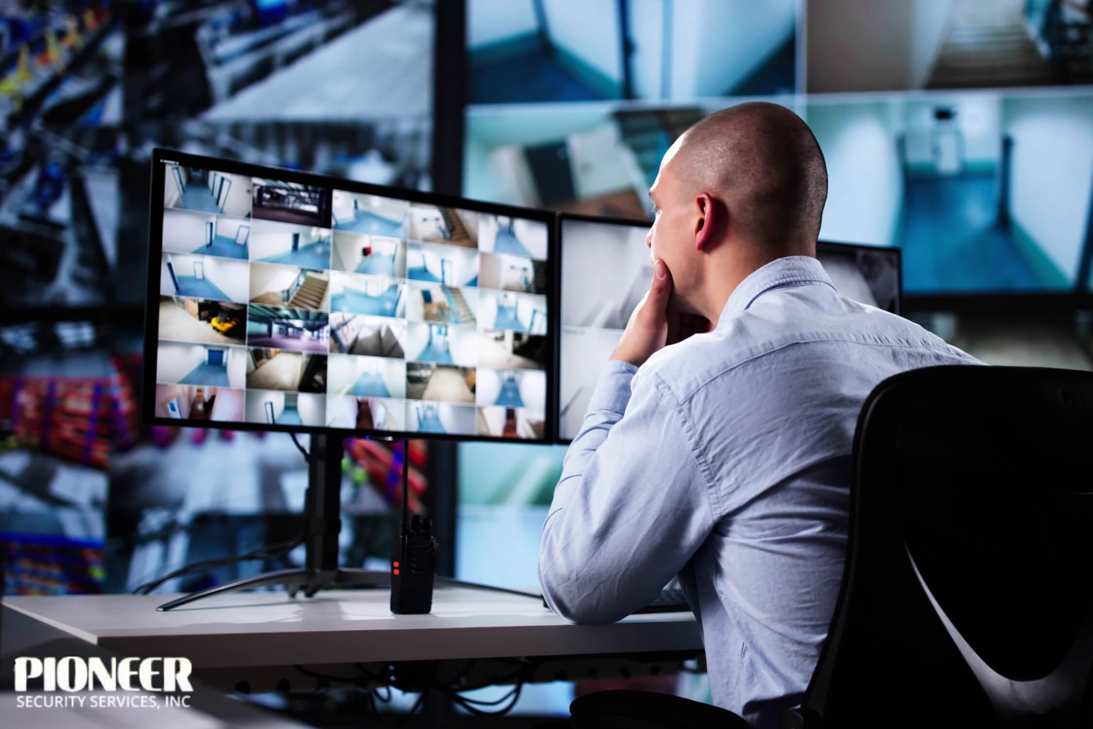 A bald man in a light blue shirt sits at a desk in a security control room, looking intently at a multi-monitor setup. One monitor displays a grid of many small surveillance video feeds, mostly showing hallways and stairwells, while a two-way radio sits on the desk in front of him.