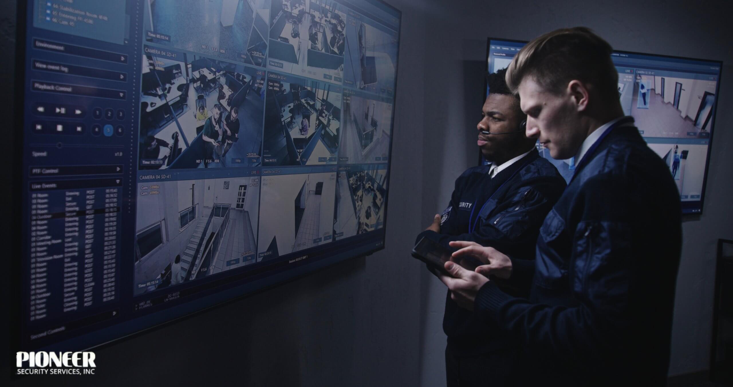 Two male security officers wearing black shirts and headsets are seated at a desk, looking intently at surveillance monitors. One officer points at a screen displaying a grid of camera feeds, while the other officer appears surprised or concerned.